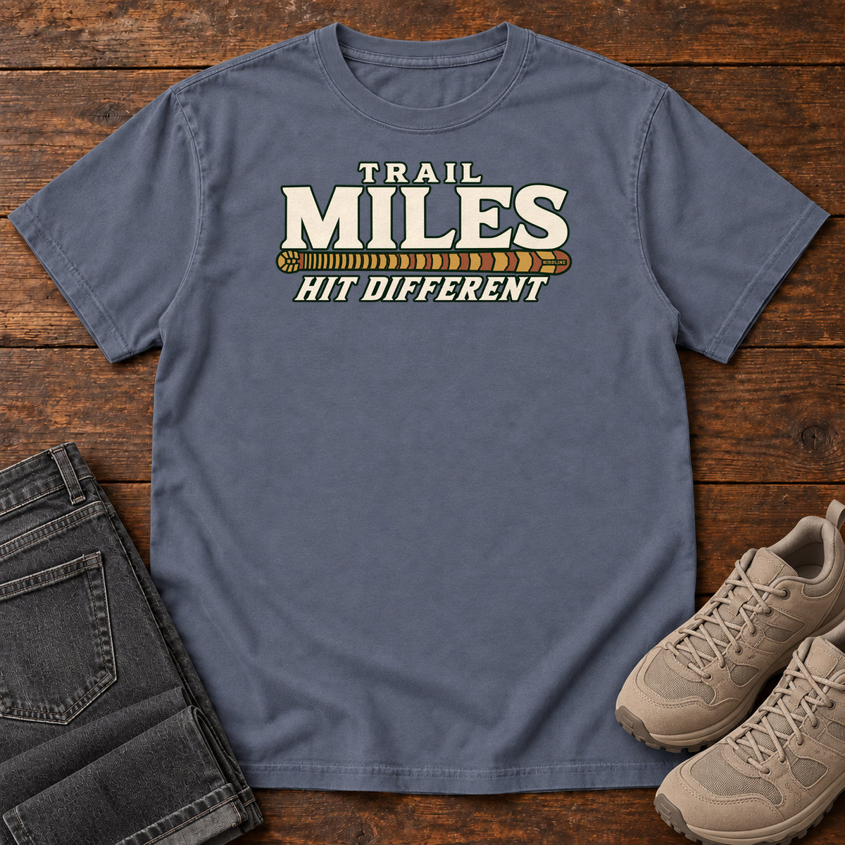 Denim Trail Miles Hit Different T-Shirt with cream lettering and a gold-to-rust patterned stripe, front flat lay on wood.