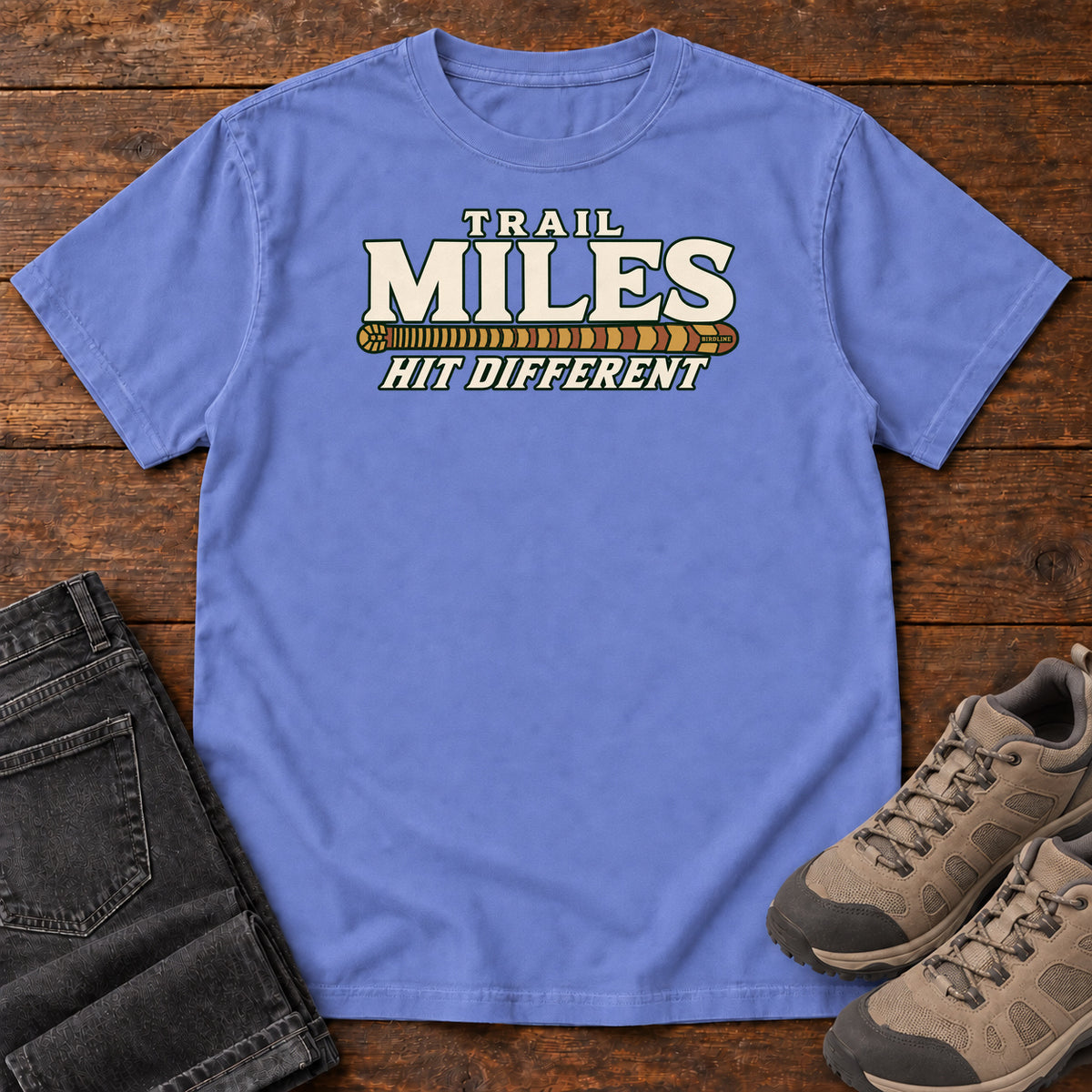 Flo Blue Trail Miles Hit Different T-Shirt with cream lettering and a gold-to-rust patterned stripe, front flat lay on wood.