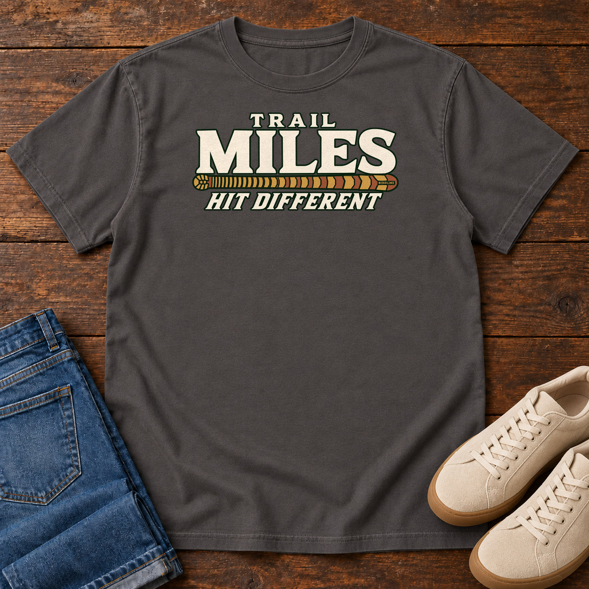 Graphite Trail Miles Hit Different T-Shirt with cream lettering and a gold-to-rust patterned stripe, front flat lay on wood.