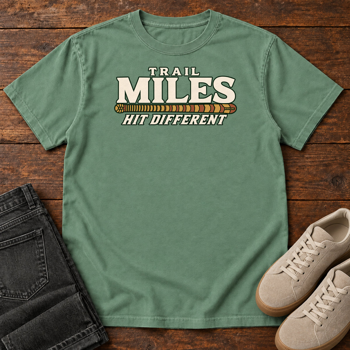 Light Green Trail Miles Hit Different T-Shirt with cream lettering and a gold-to-rust patterned stripe, front flat lay on wood.