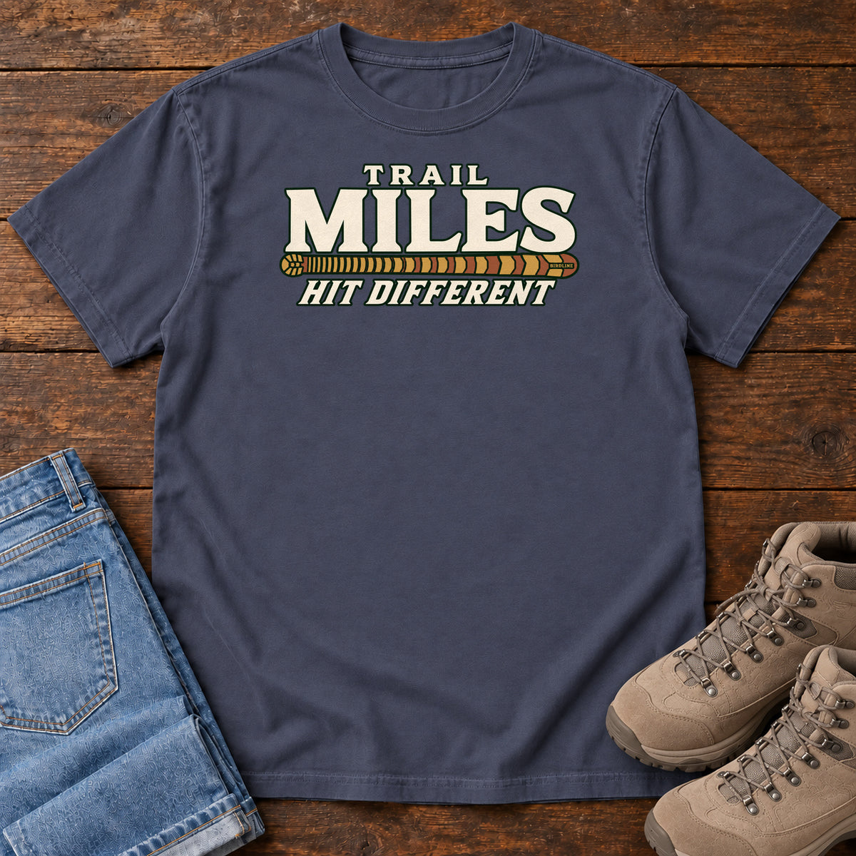 Navy Trail Miles Hit Different T-Shirt with cream lettering and a gold-to-rust patterned stripe, front flat lay on wood.