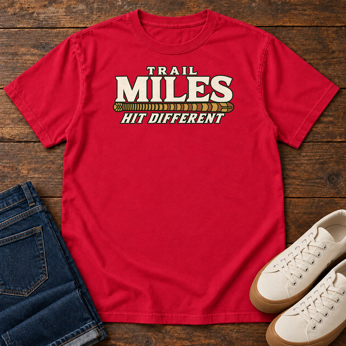 Red Trail Miles Hit Different T-Shirt with cream lettering and a gold-to-rust patterned stripe, front flat lay on wood.