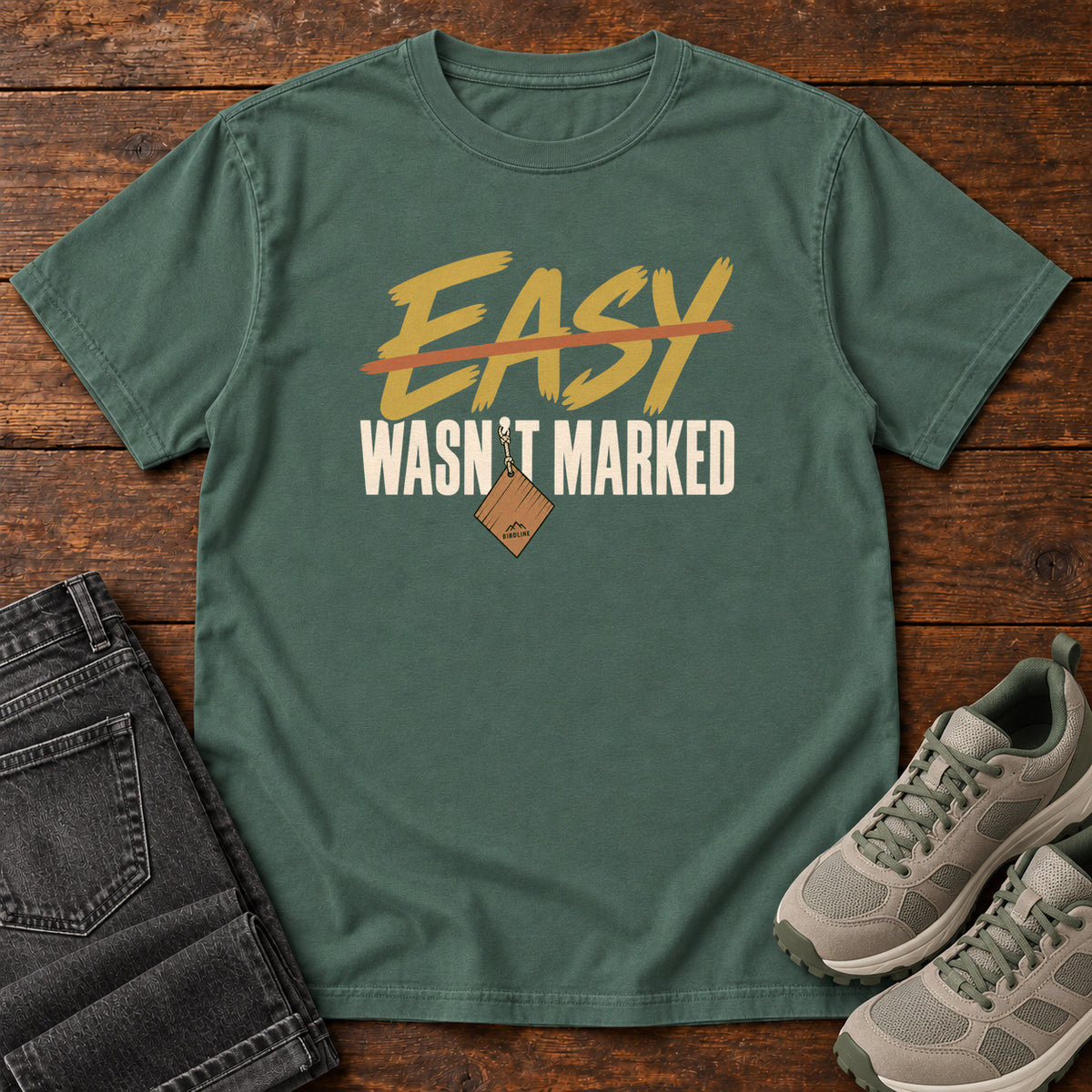 Blue Spruce Easy Wasn't Marked T-Shirt with gold Easy lettering crossed out in orange above cream Wasn't Marked text, front flat lay on wood.