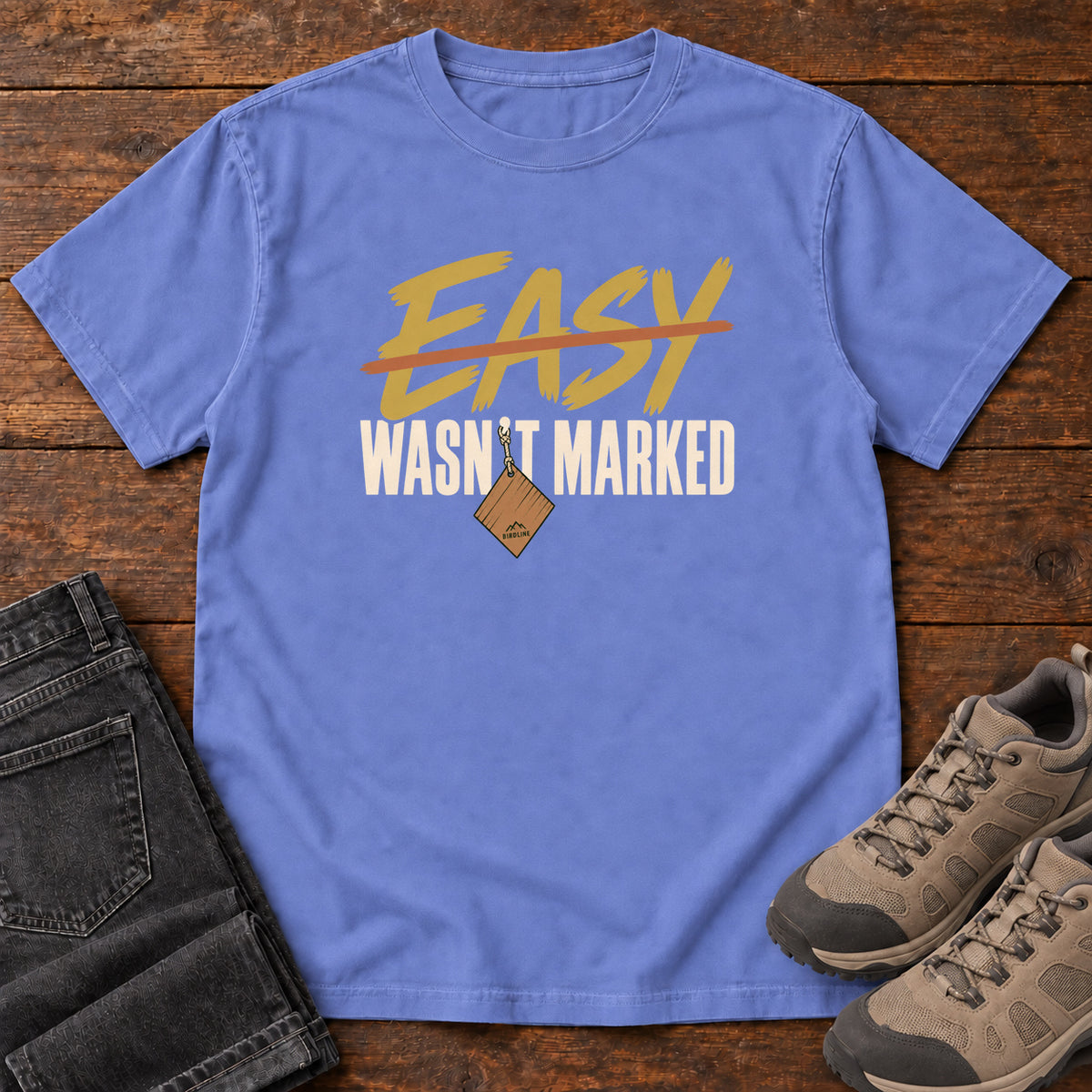 Flo Blue Easy Wasn't Marked T-Shirt with gold Easy lettering crossed out in orange above cream Wasn't Marked text, front flat lay on wood.