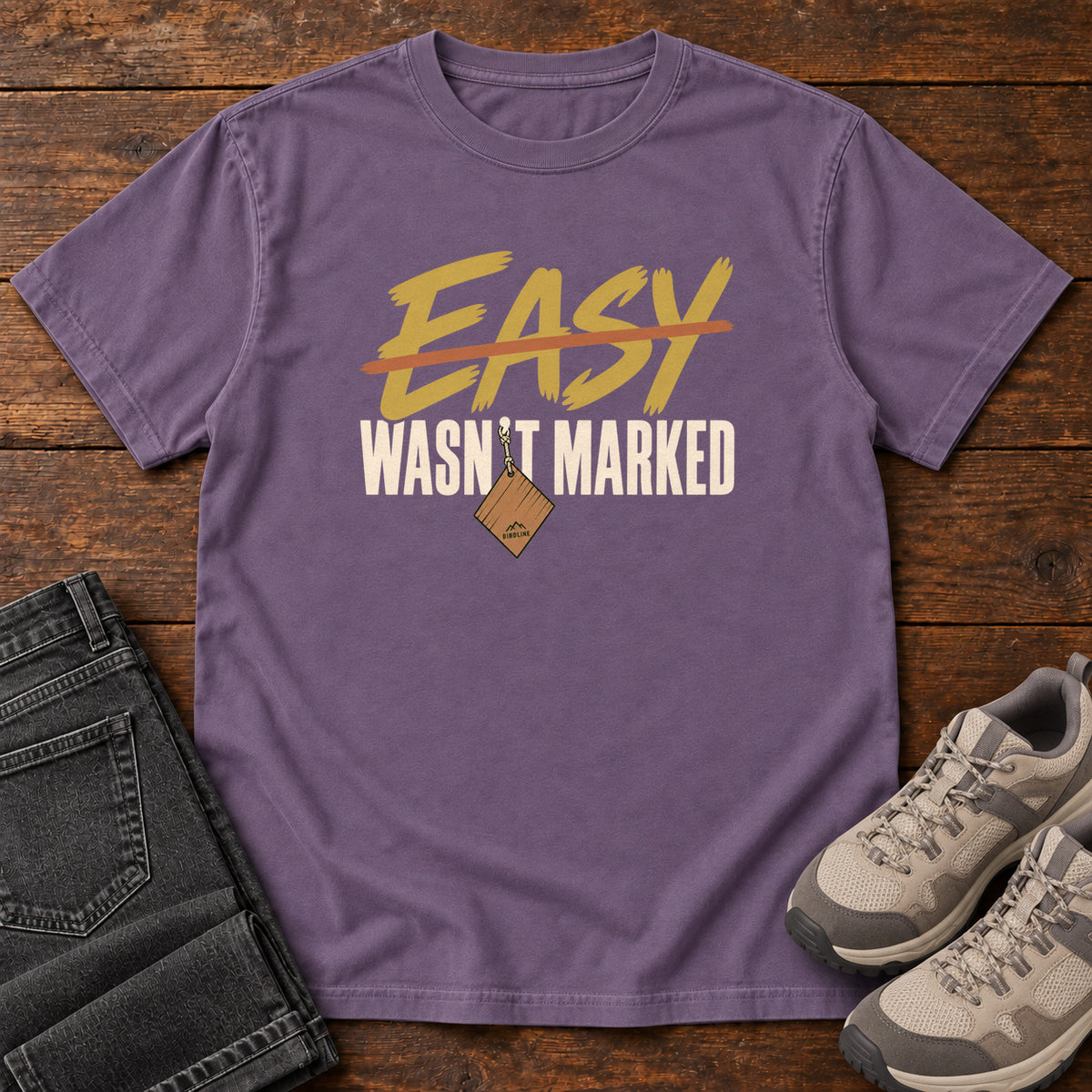 Grape Easy Wasn't Marked T-Shirt with gold Easy lettering crossed out in orange above cream Wasn't Marked text, front flat lay on wood.