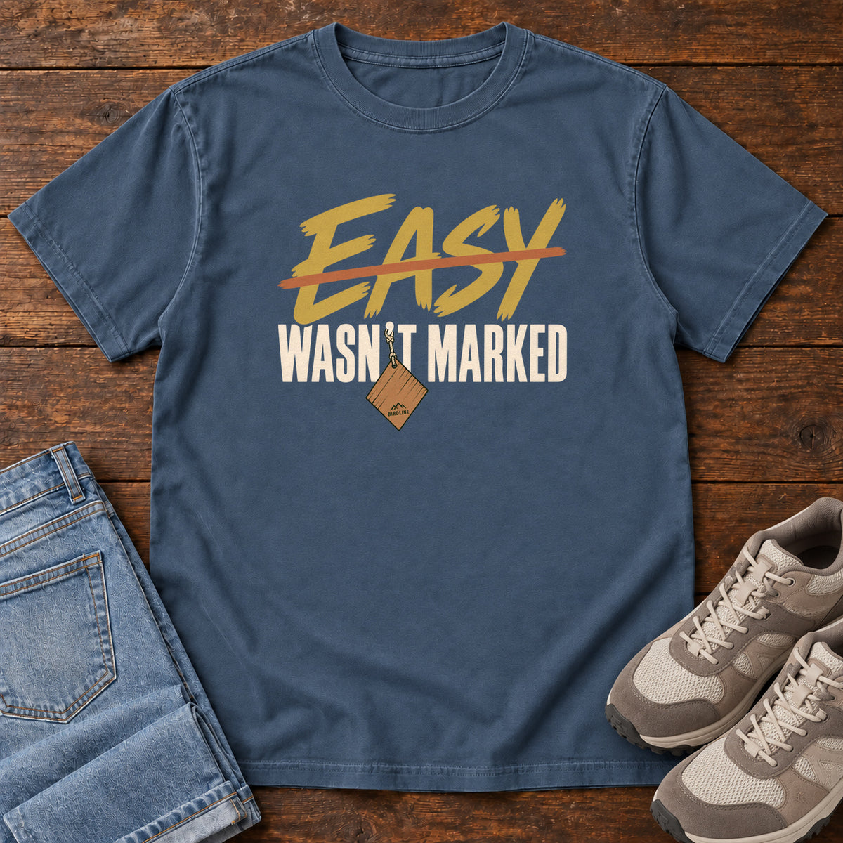 Midnight Easy Wasn't Marked T-Shirt with gold Easy lettering crossed out in orange above cream Wasn't Marked text, front flat lay on wood.