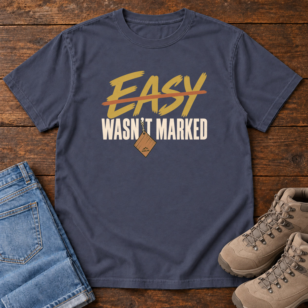 Navy Easy Wasn't Marked T-Shirt with gold Easy lettering crossed out in orange above cream Wasn't Marked text, front flat lay on wood.