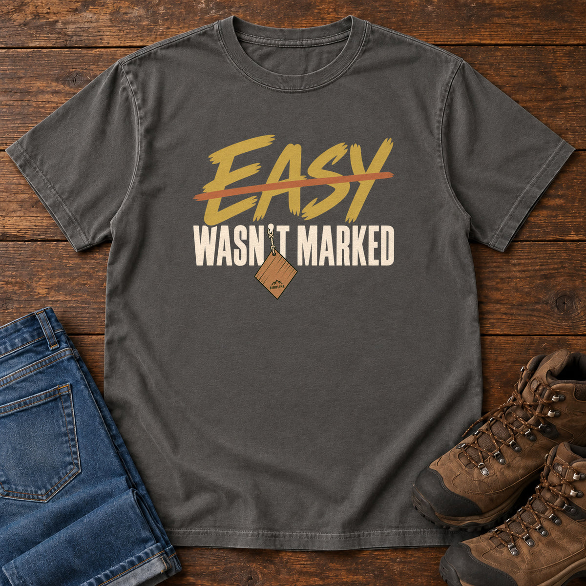 Pepper Easy Wasn't Marked T-Shirt with gold Easy lettering crossed out in orange above cream Wasn't Marked text, front flat lay on wood.
