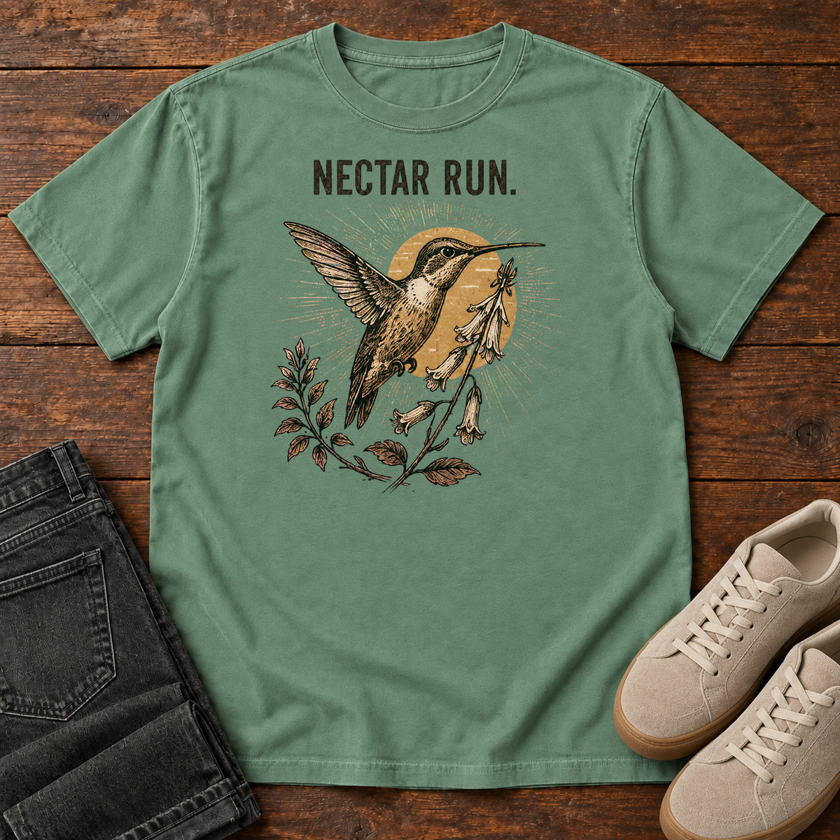 Light Green Nectar Run Sunburst T-Shirt with a long-beaked bird hovering beside flowers against a golden sunburst; front flat lay on wood.