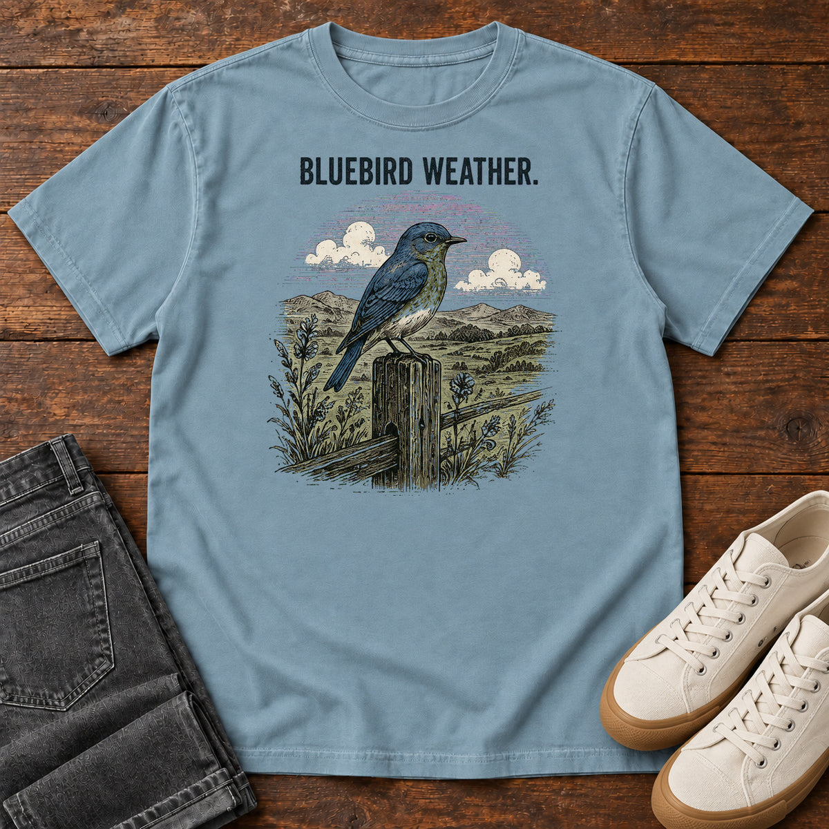 Ice Blue Bluebird Weather Meadow T-Shirt with a blue-and-orange bird on a fence post overlooking flowers, hills and clouds; front flat lay on wood.