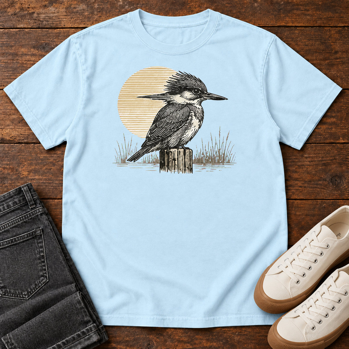 Chambray Kingfisher At Rest T-Shirt with a crested black-and-white bird perched on a post in front of a pale sun, front flat lay on wood.