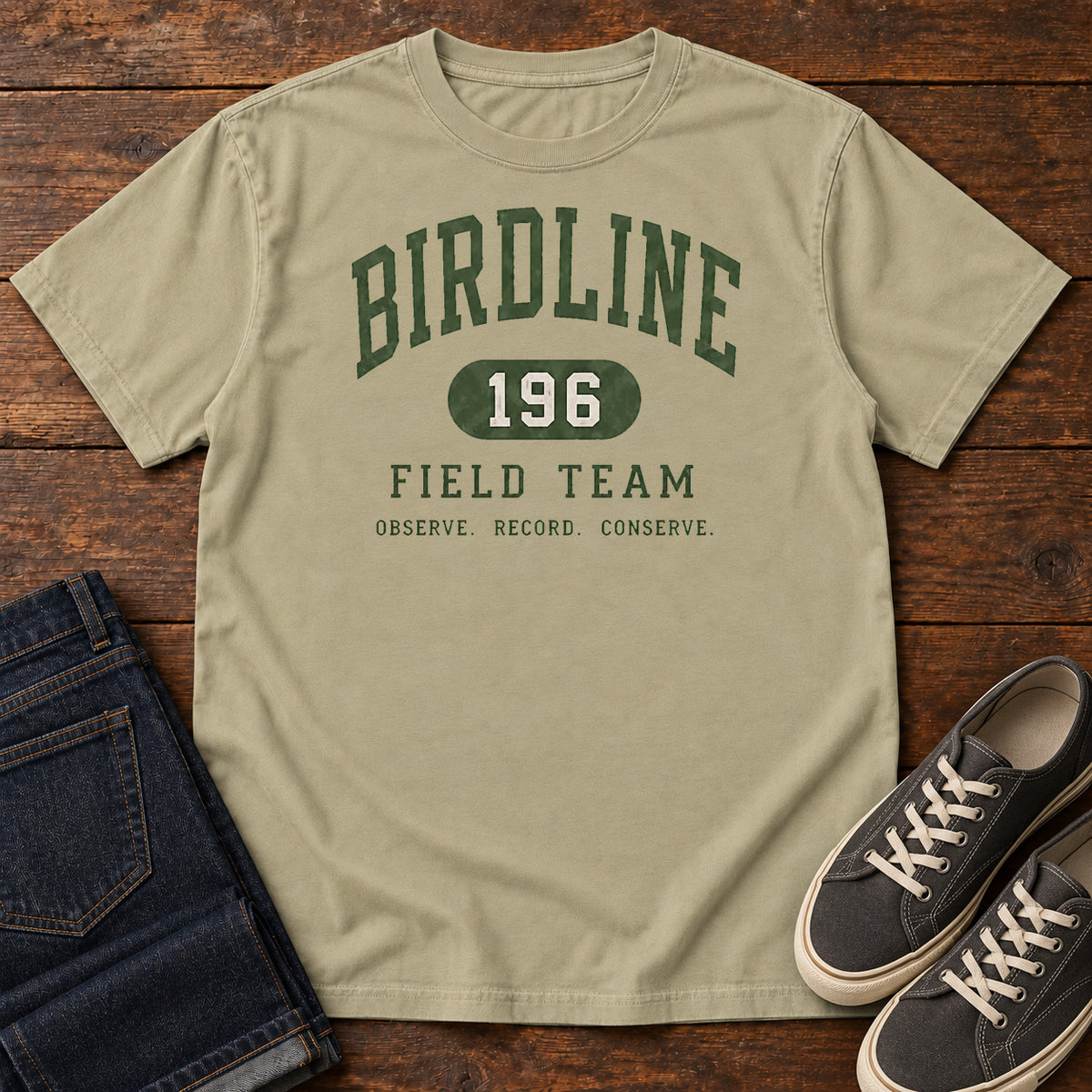 Khaki Birdline Field Team T-Shirt with green collegiate Birdline Field Team lettering and the number 196, front flat lay on wood.