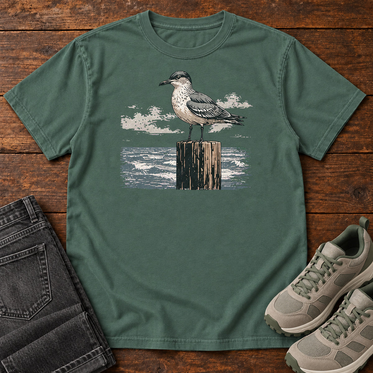 Blue Spruce Gull On A Piling T-shirt with a gull standing on a wooden piling above the water, laid flat on a wooden surface.