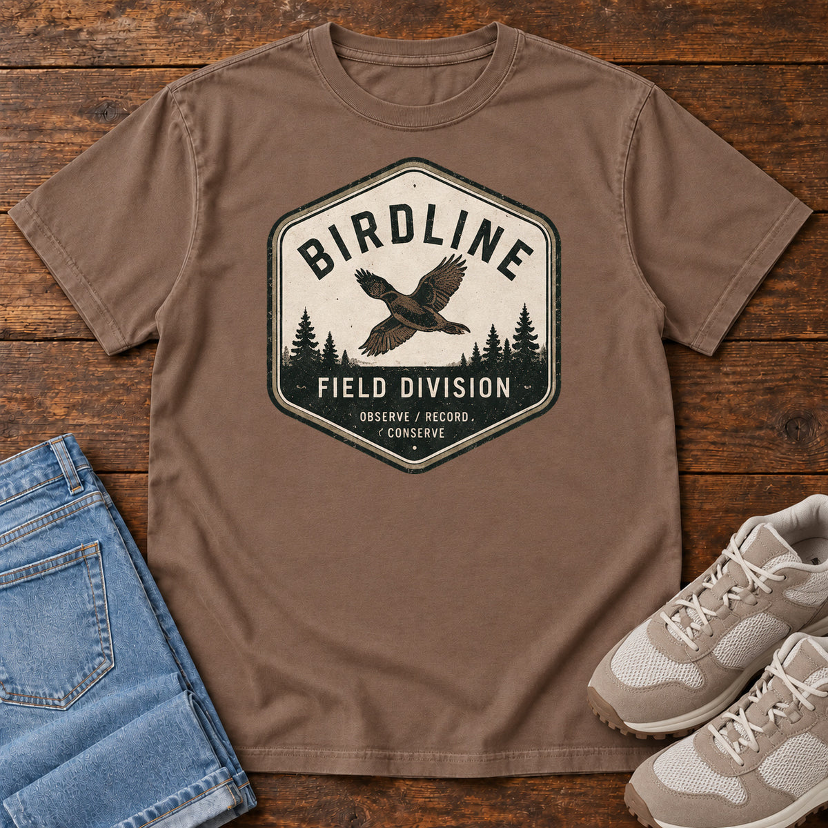 Espresso Birdline Field Division T-Shirt with a cream badge showing a flying bird over pine trees; front flat lay on wood.