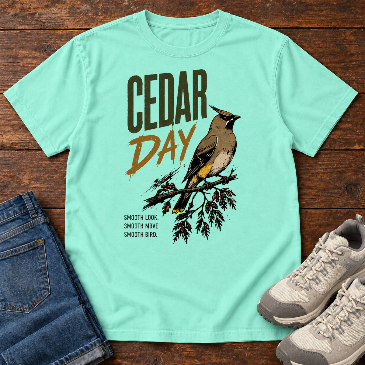 Chalky Mint Cedar Day T-shirt with a crested bird on a branch and Cedar Day lettering, laid flat on a wooden surface.