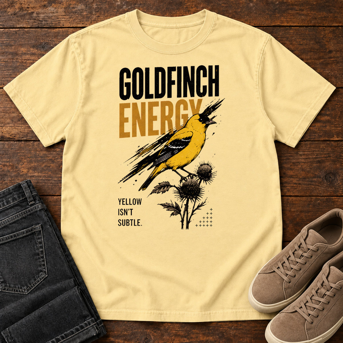 Butter Goldfinch Energy T-shirt with a yellow bird on thistles and Goldfinch Energy lettering, laid flat on a wooden surface.