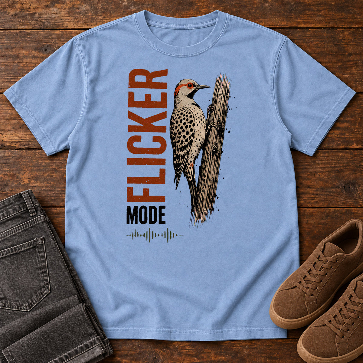 Washed Denim Flicker Mode T-shirt with a spotted bird on a tree trunk and Flicker Mode lettering, laid flat on a wooden surface.