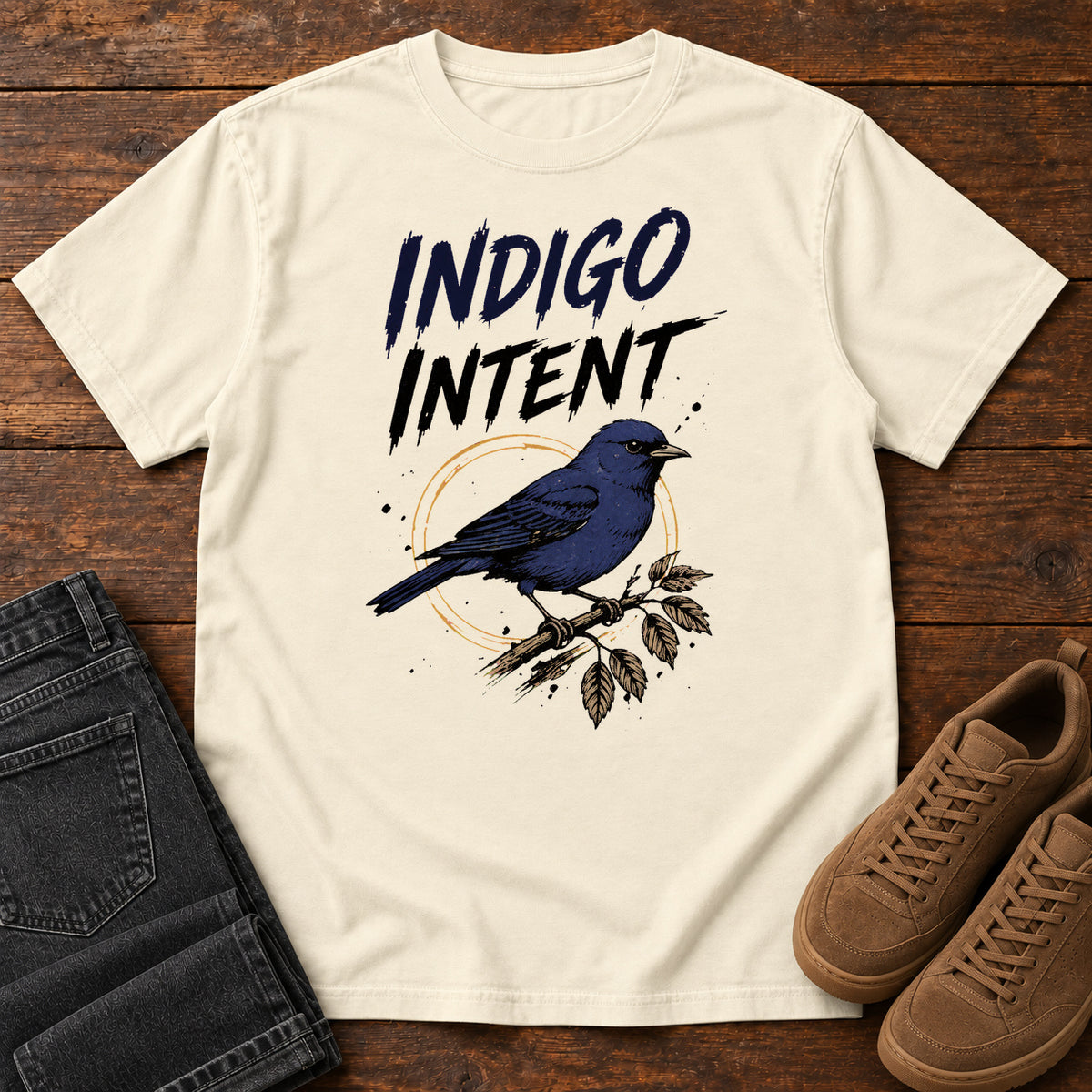 Ivory Indigo Intent T-shirt with a blue bird on a branch and Indigo Intent lettering, laid flat on a wooden surface.