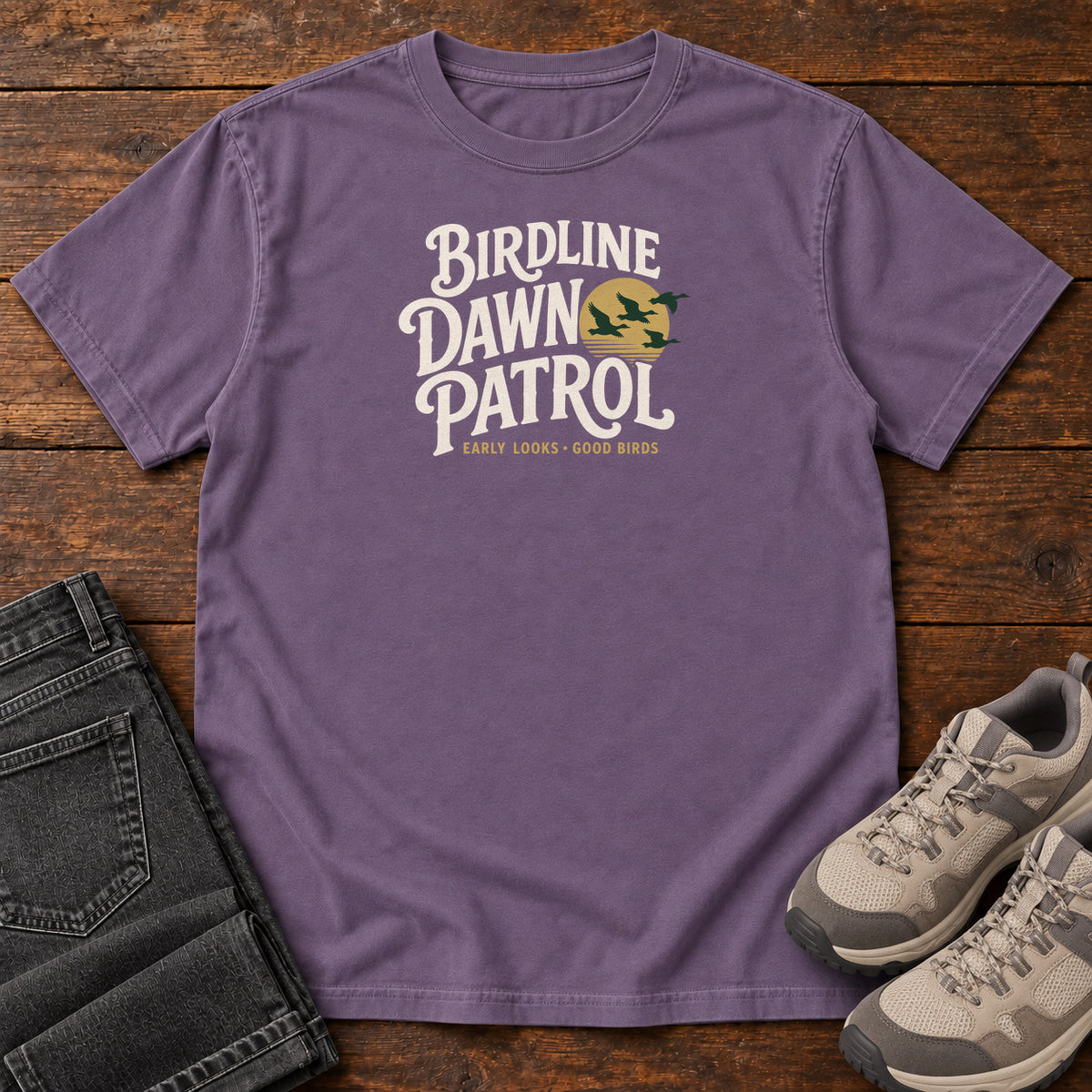 Birdline Dawn Patrol T-shirt in Grape, front laid flat on wood, featuring cream BIRDLINE DAWN PATROL lettering beside dark flying birds against a gold sun.
