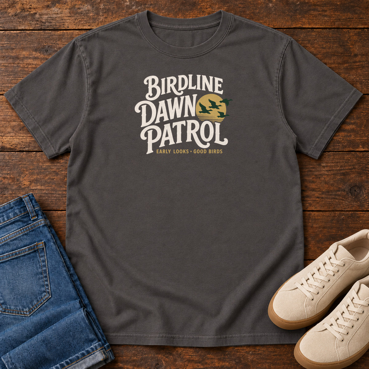 Birdline Dawn Patrol T-shirt in Graphite, front laid flat on wood, featuring cream BIRDLINE DAWN PATROL lettering beside dark flying birds against a gold sun.
