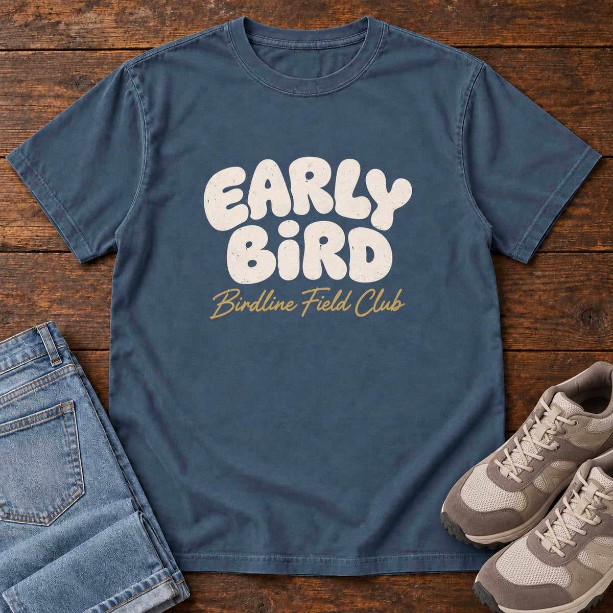 Midnight Early Bird Field Club T-shirt with cream bubble lettering with a gold script subtitle; front flat lay on wood.