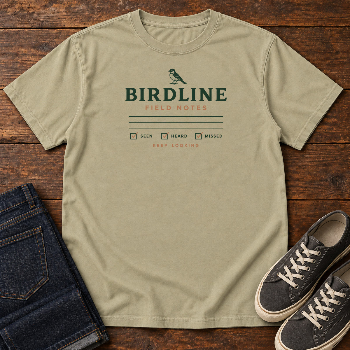 Khaki Field Notes T-shirt with a small bird above Birdline Field Notes lettering, ruled lines and checkboxes, front flat lay on wood.