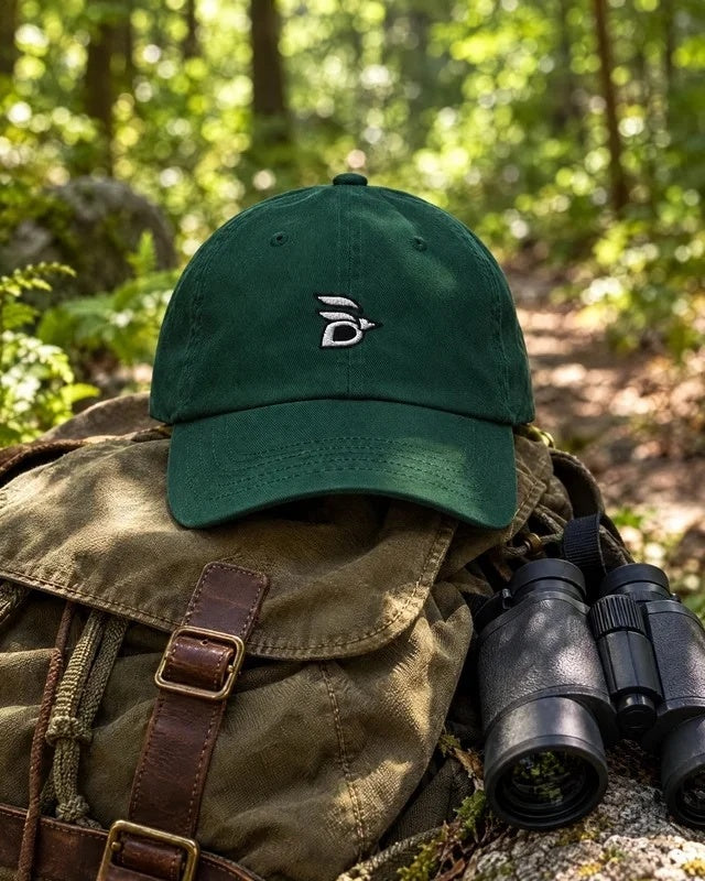 Birdline Logo Dad Hat in Spruce on a canvas backpack with binoculars on a forest trail