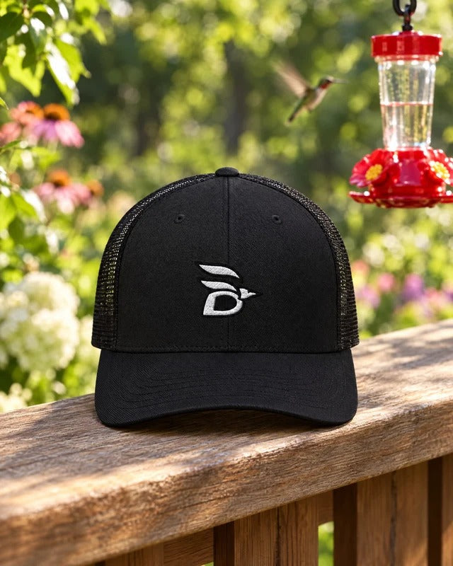 Birdline Logo Trucker Hat in Black on a porch rail beside a hummingbird feeder