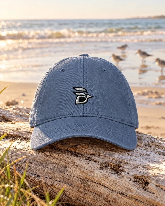Birdline Logo Washed Cap in Blue Jean on driftwood at the beach, with sandpipers behind