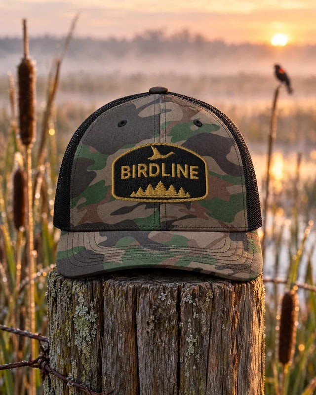 Birdline Pinewood Flight Camo Trucker Hat in Camo/Black on a fence post in a misty cattail marsh at sunrise